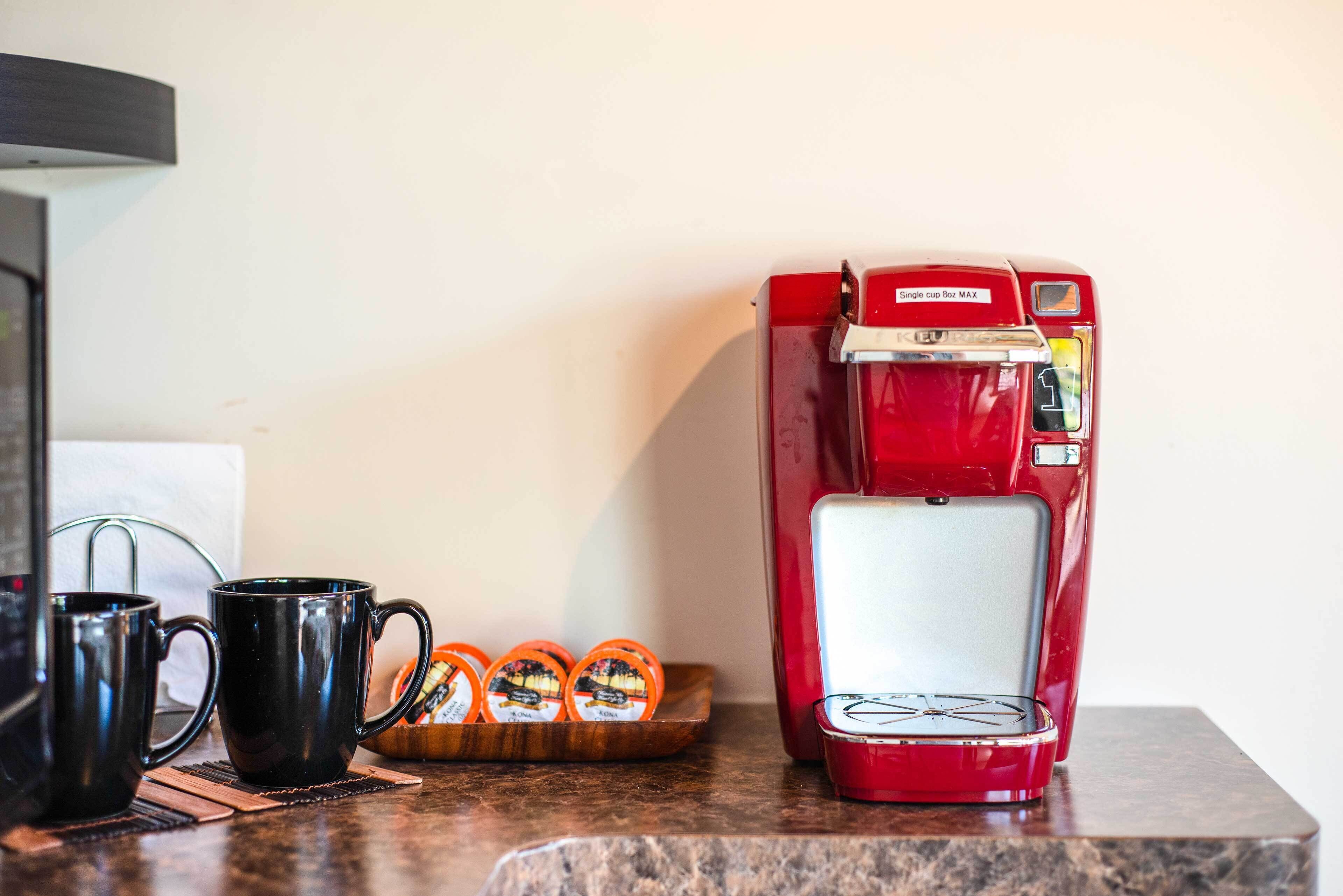 Une machine à café rouge est posée sur un plan de travail de cuisine, à côté de deux tasses noires et d’un plateau de dosettes de café.