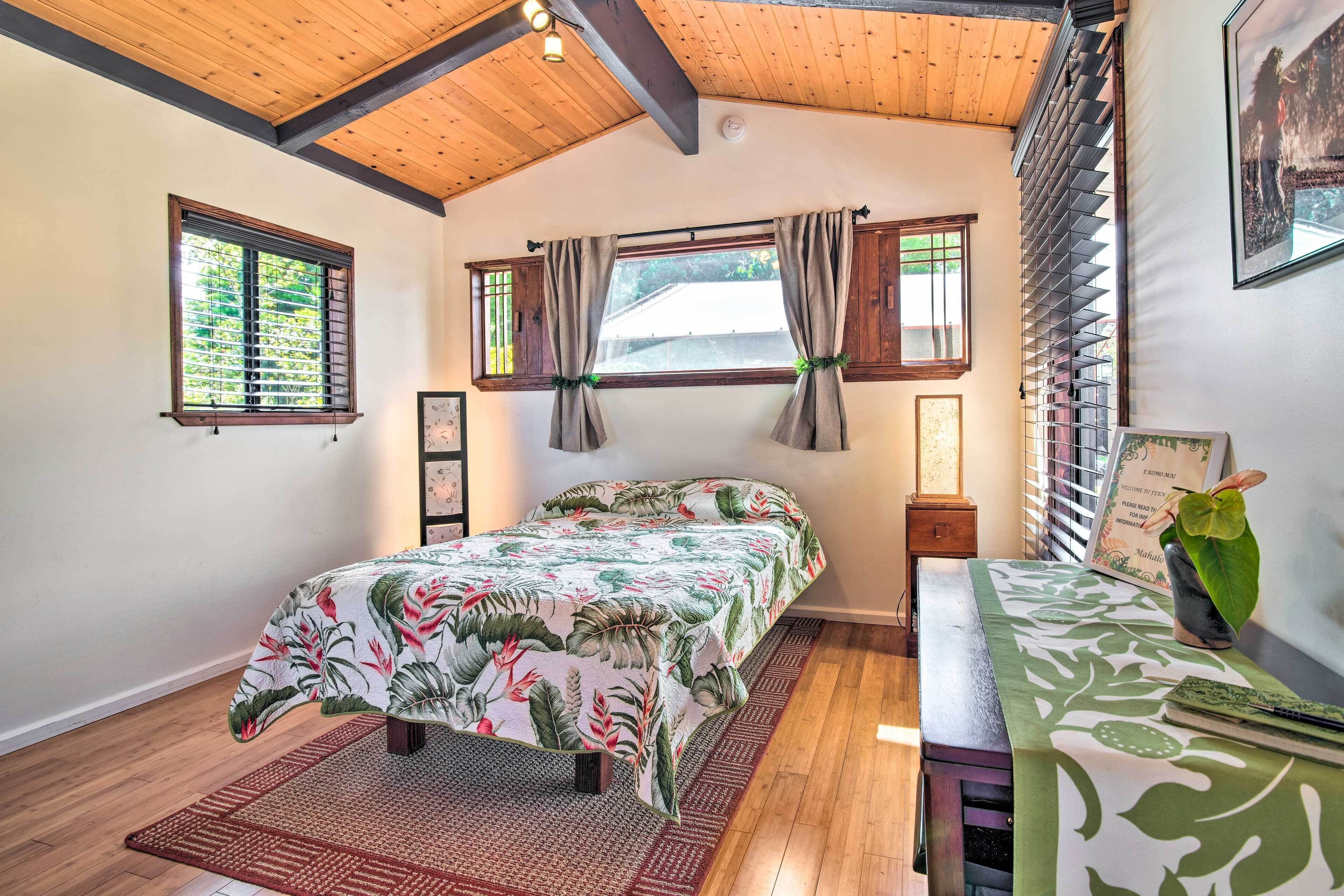 A bedroom with a bed, a nightstand, a window with curtains, a wooden ceiling, and a wall-mounted lamp.