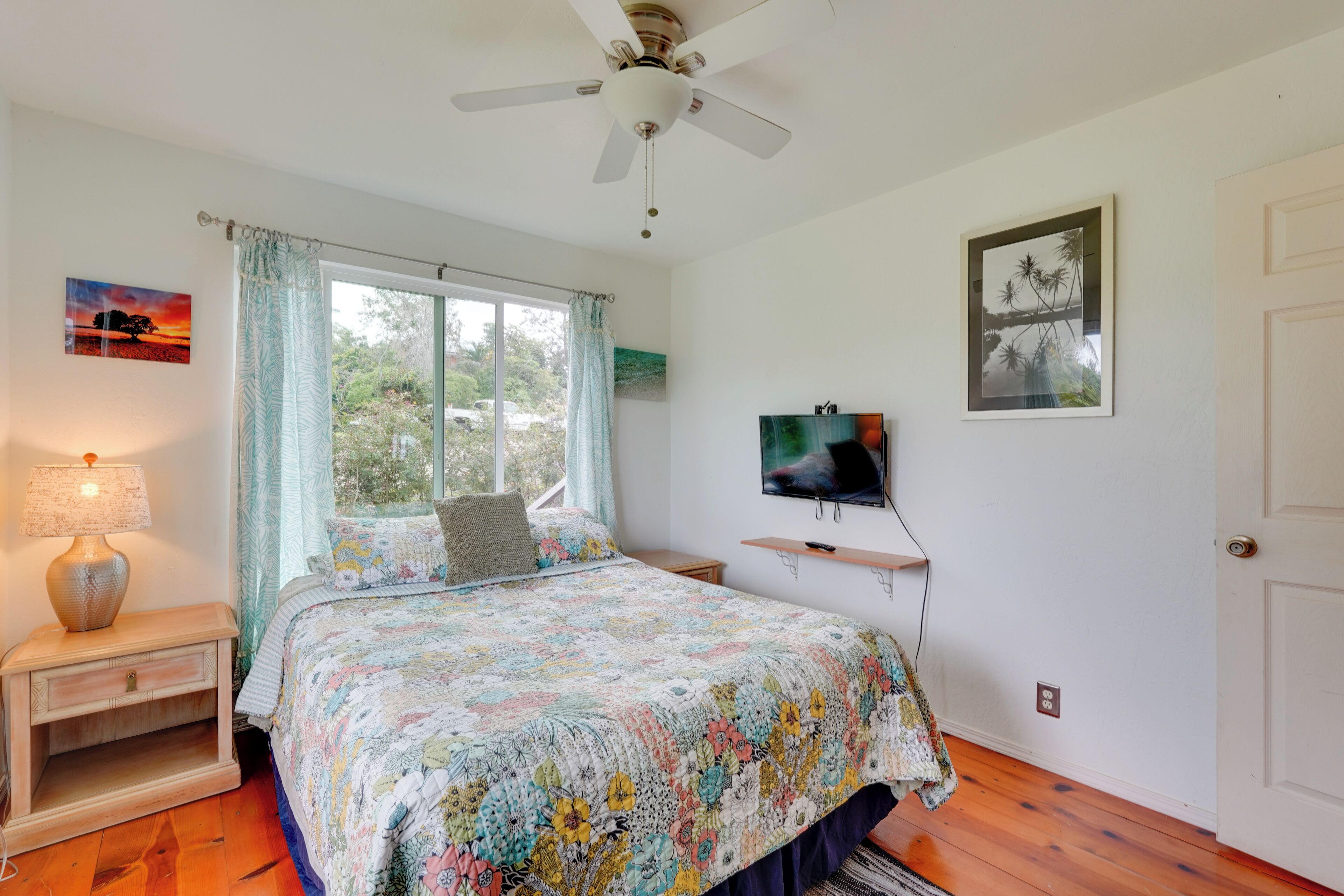 Un dormitorio con cama, ventilador de techo, televisión y ventana con cortinas.
