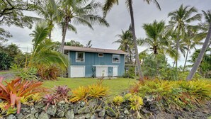 A two-story house with a blue exterior, surrounded by tropical vegetation and palm trees. - Breezy Kailua-kona Bungalow w/ Lanai & Ocean View! (Kailua-Kona)