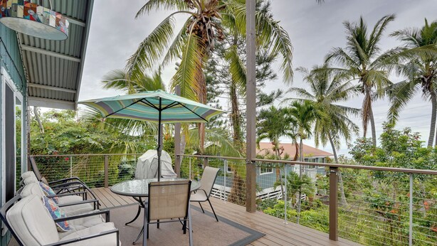A wooden deck with outdoor furniture, including chairs and a table, under a striped umbrella. - Breezy Kailua-kona Bungalow w/ Lanai & Ocean View! (Kailua-Kona)