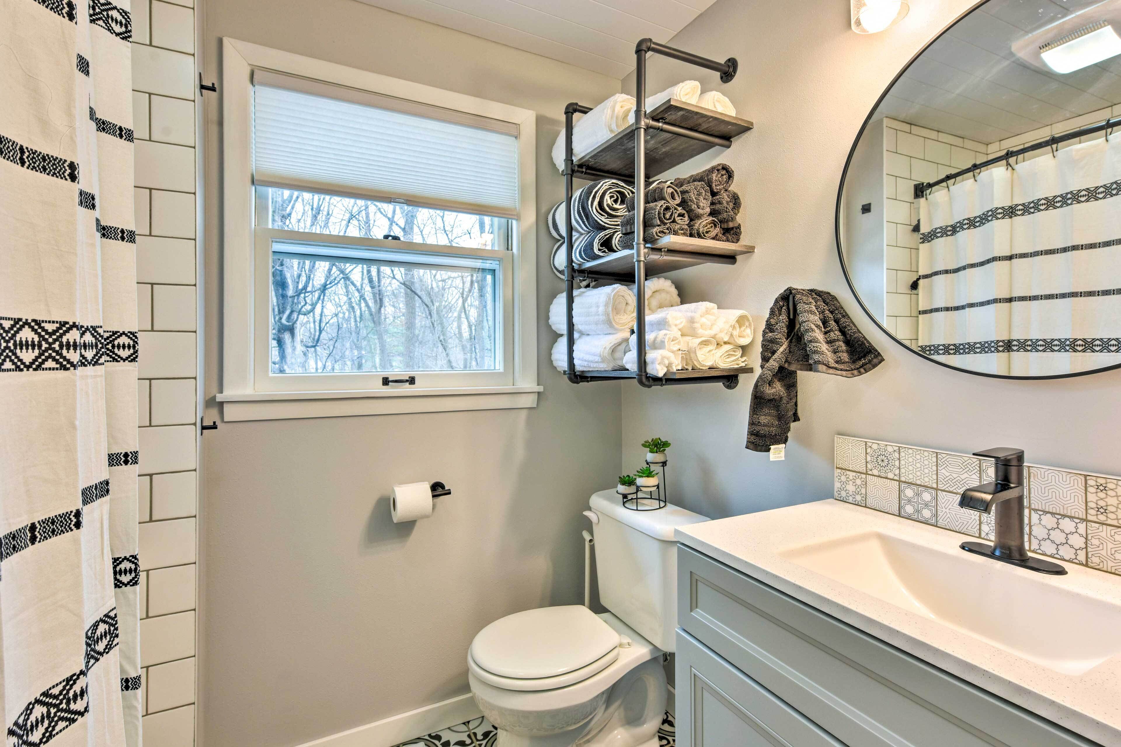 A bathroom with a toilet, a sink with a modern faucet, a towel rack with folded towels, a window with blinds, and a patterned tile backsplash.