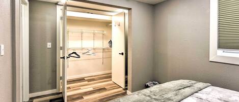 A bedroom with a bed, a gray blanket, a wooden floor, a closet with a mirror, and a window with blinds.