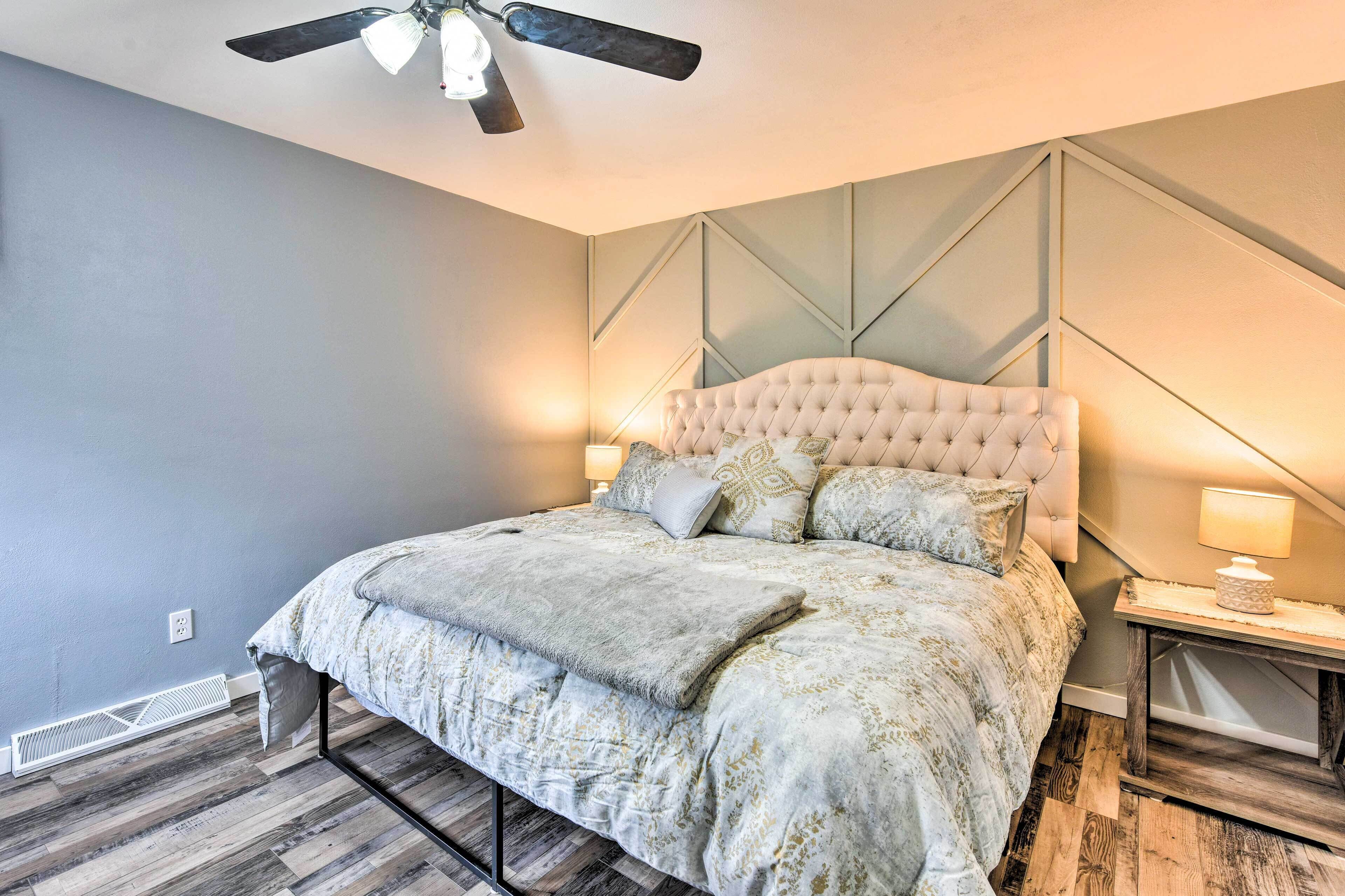 A bedroom with a tufted headboard, a bed with a patterned duvet, a wooden floor, and a ceiling fan.