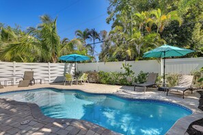 A swimming pool with a tiled edge, surrounded by lounge chairs and umbrellas, with palm trees and a white fence in the background. - Siesta Key Dream Inn Vacation Rental Near Village! (Siesta Key)