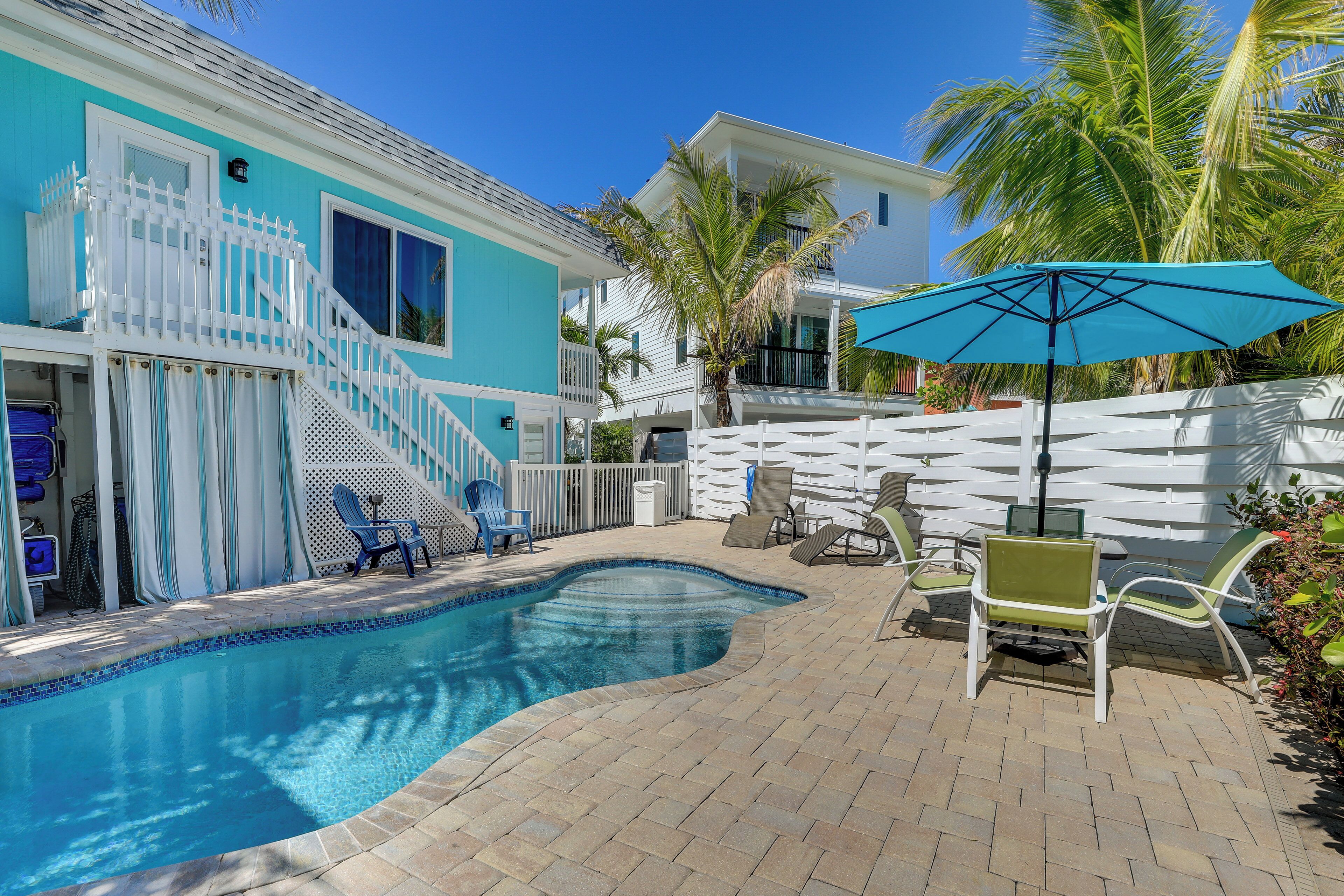 A pool area with a white fence, a blue umbrella, and lounge chairs.