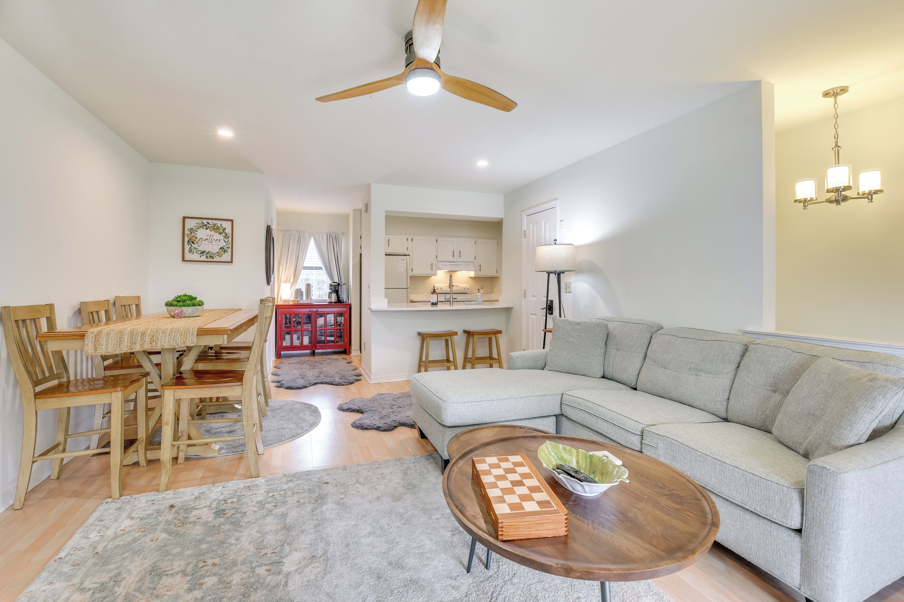 A spacious living room with a large sectional sofa, a wooden coffee table with a chess set, a dining area with a wooden table and chairs, and a kitchen in the background.