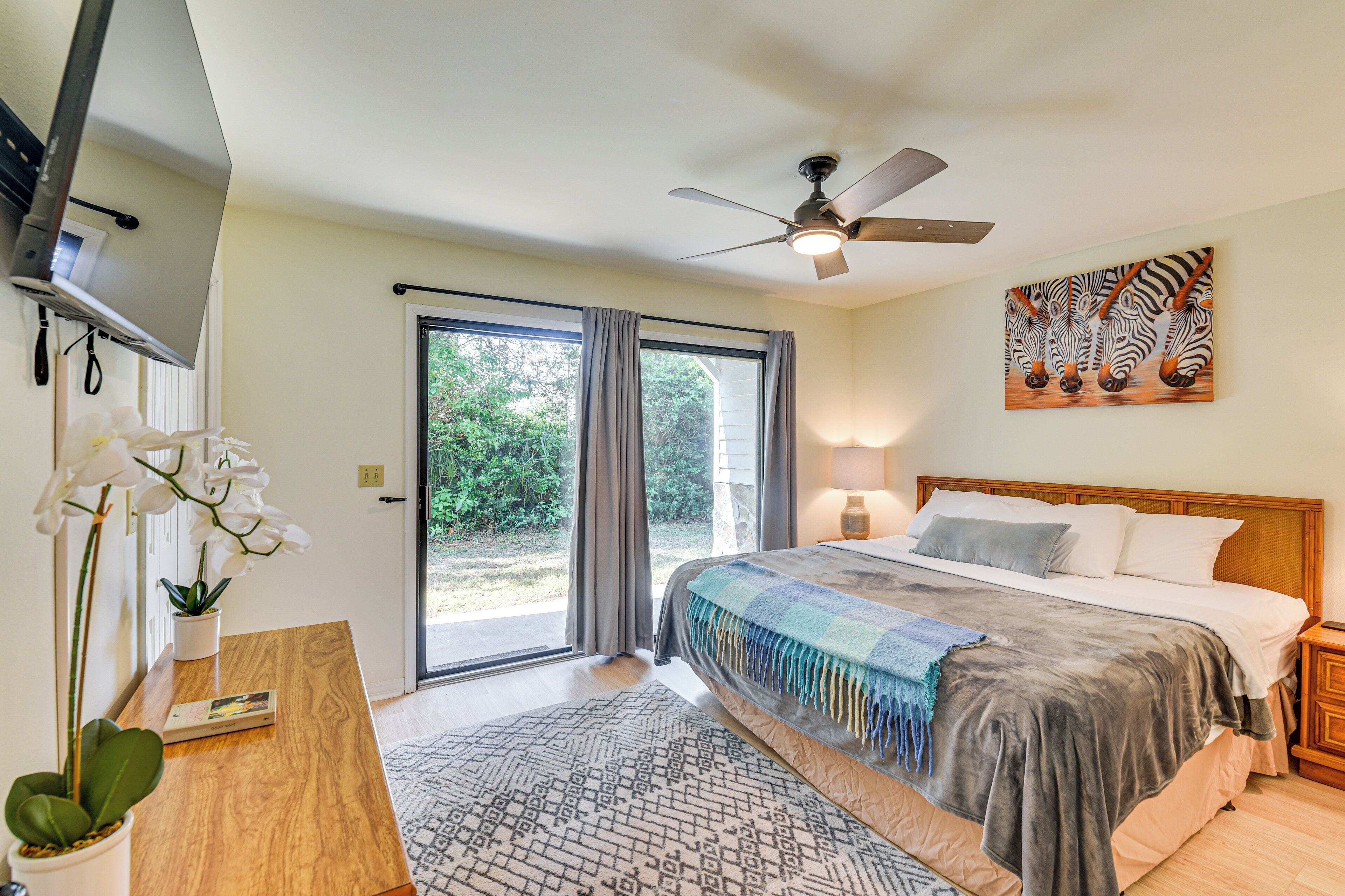 Une chambre à coucher avec un lit, un ventilateur de plafond, un tableau représentant des zèbres et une vue sur la verdure à l’extérieur.