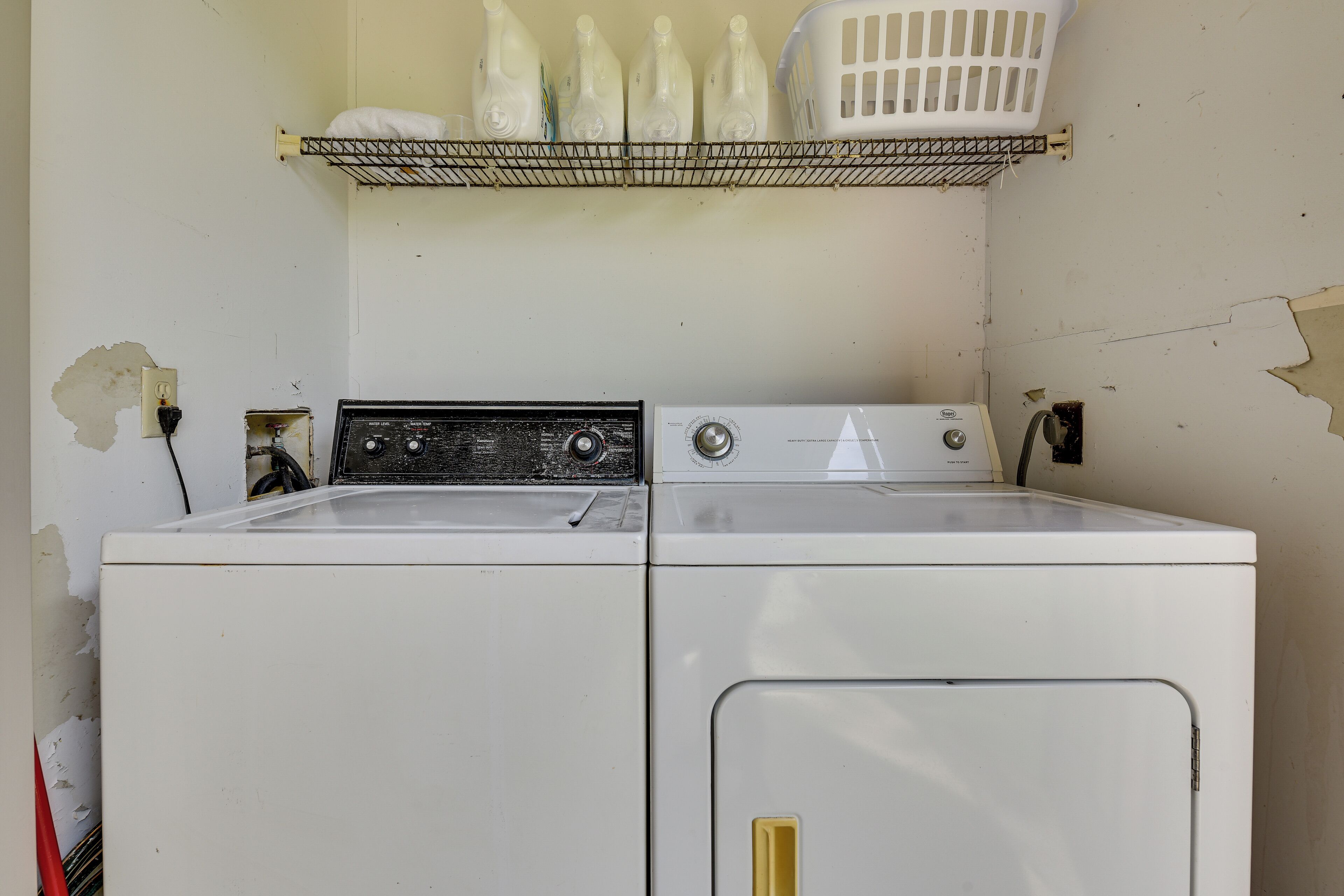 A laundry area with two washing machines, a shelf with laundry baskets, and a damaged wall.