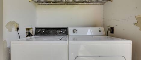 A laundry area with two washing machines, a shelf with laundry baskets, and a damaged wall.