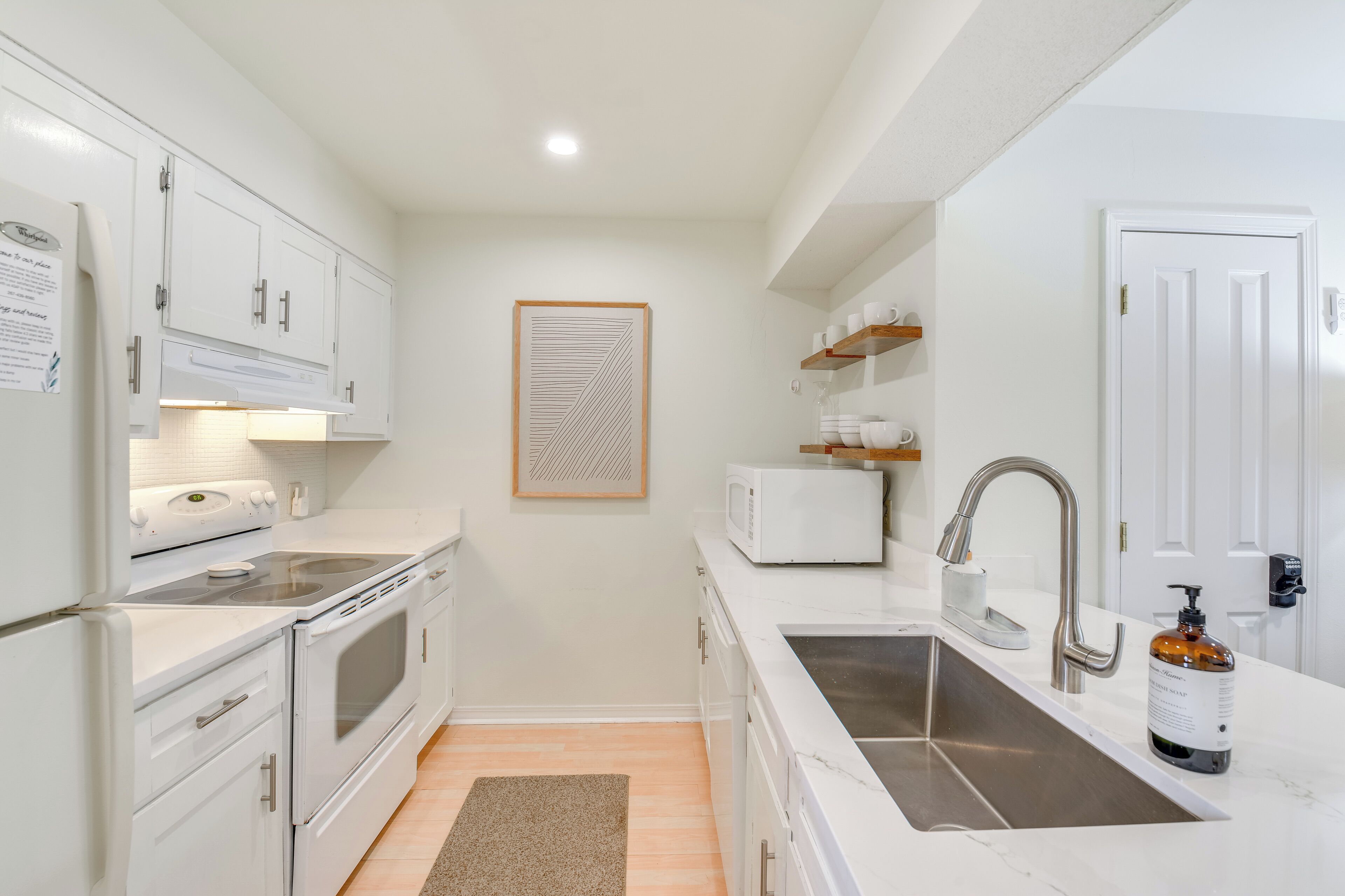 Una cocina compacta con muebles blancos, un lavamanos de acero inoxidable, un microondas y un refrigerador.