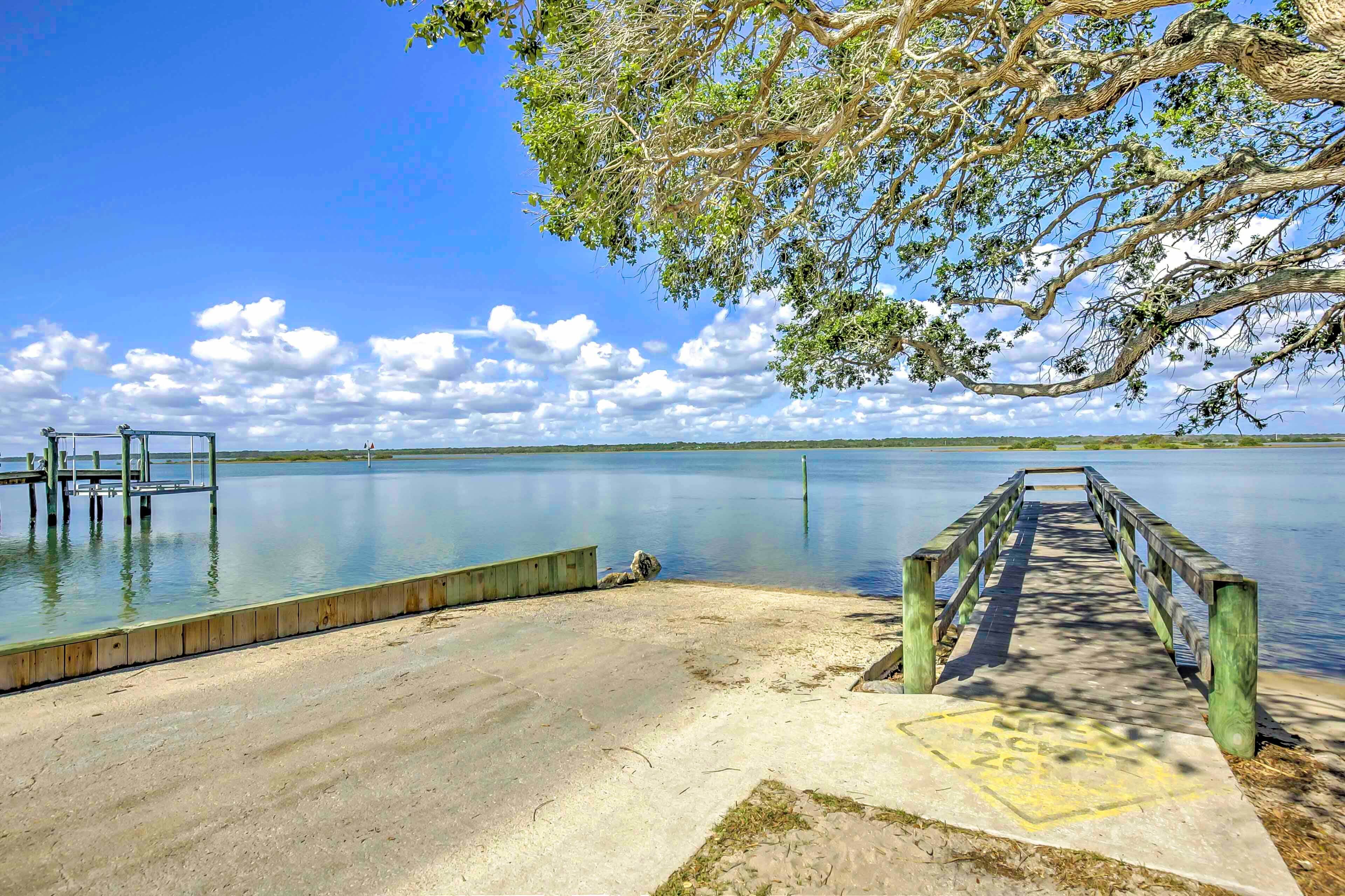 A wooden dock extending into calm water, surrounded by a sandy beach and a large tree.