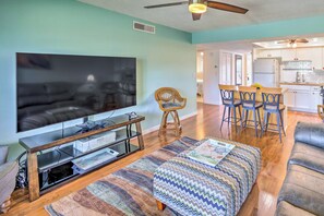 A living room with a TV, ottoman, and chairs, a kitchen with white cabinets, and a ceiling fan. - Sunny St Augustine Condo w/ Community Pool! (St. Augustine)