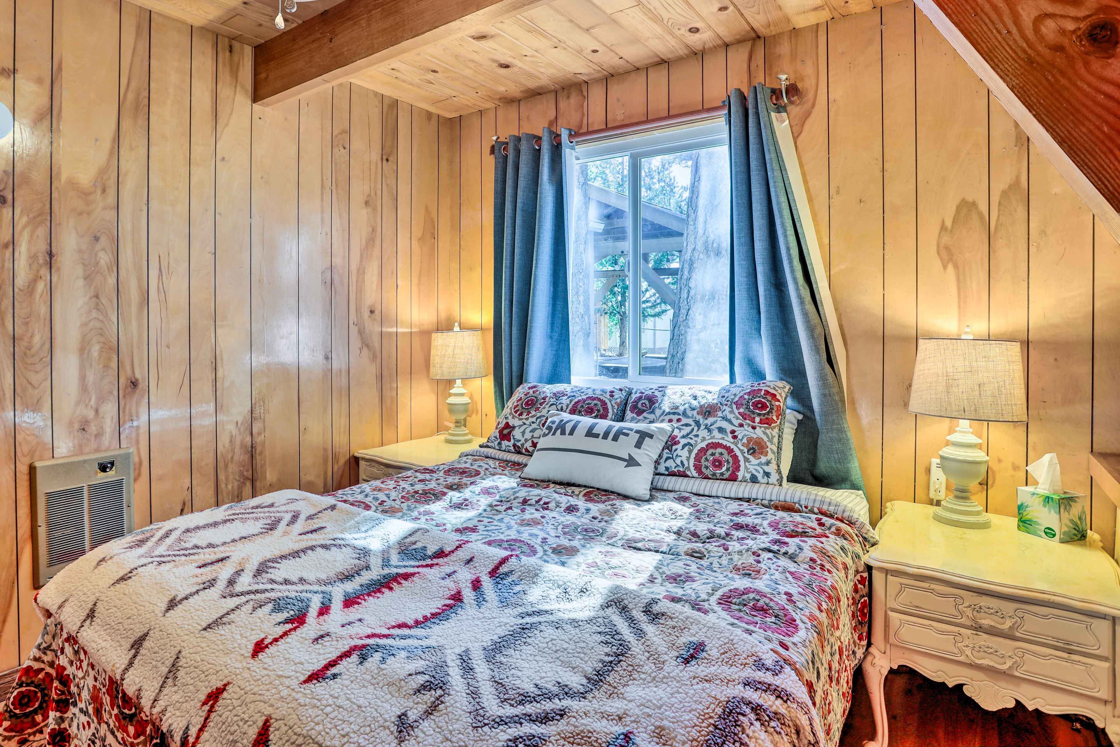 A cozy bedroom with a wooden ceiling, a bed with a patterned blanket, a nightstand with a lamp, and a window with curtains.