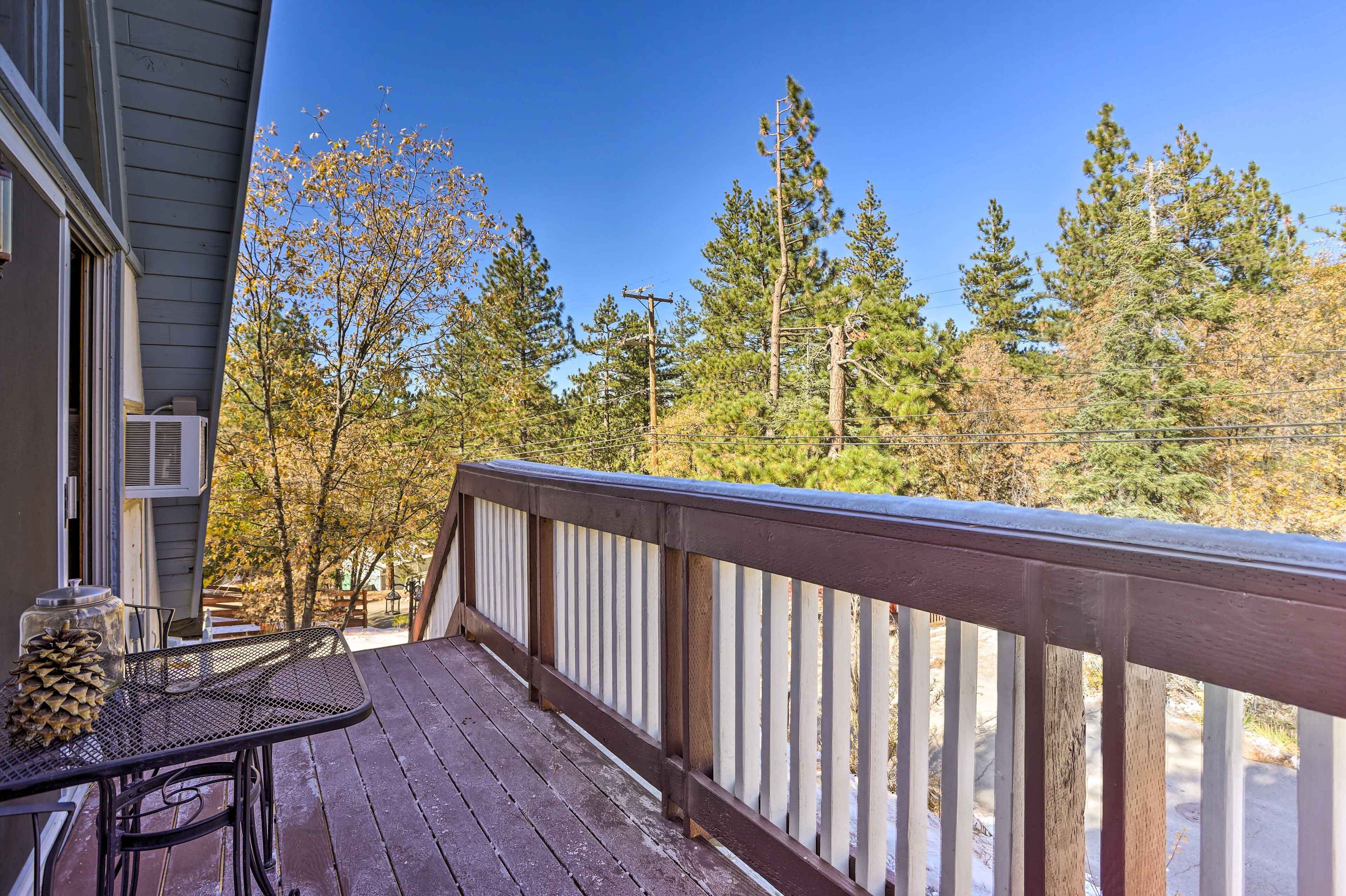 Une terrasse en bois avec une rambarde en métal, donnant sur une zone boisée où s’élèvent de grands arbres, sous un ciel bleu dégagé.