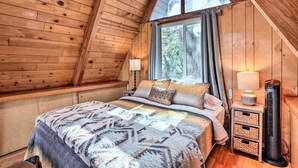 A wooden cabin bedroom with a bed, two bedside lamps, a heater, and a window with curtains. - Cozy A-frame w/ Hot Tub Near Arrowbear Lake! (Running Springs)