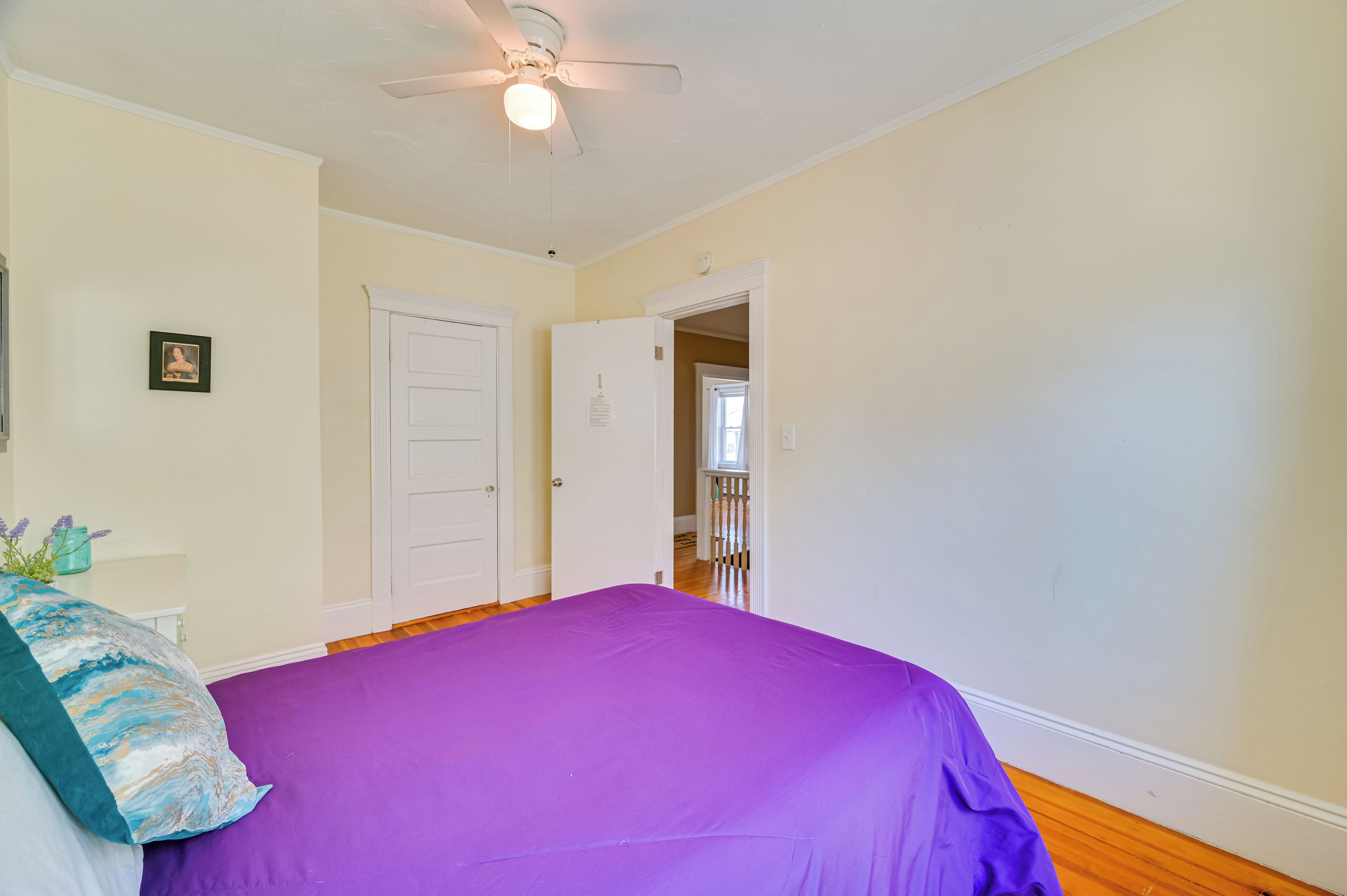 Une chambre avec une couvre-lit violette, un ventilateur de plafond, une porte blanche et un petit tableau accroché au mur.