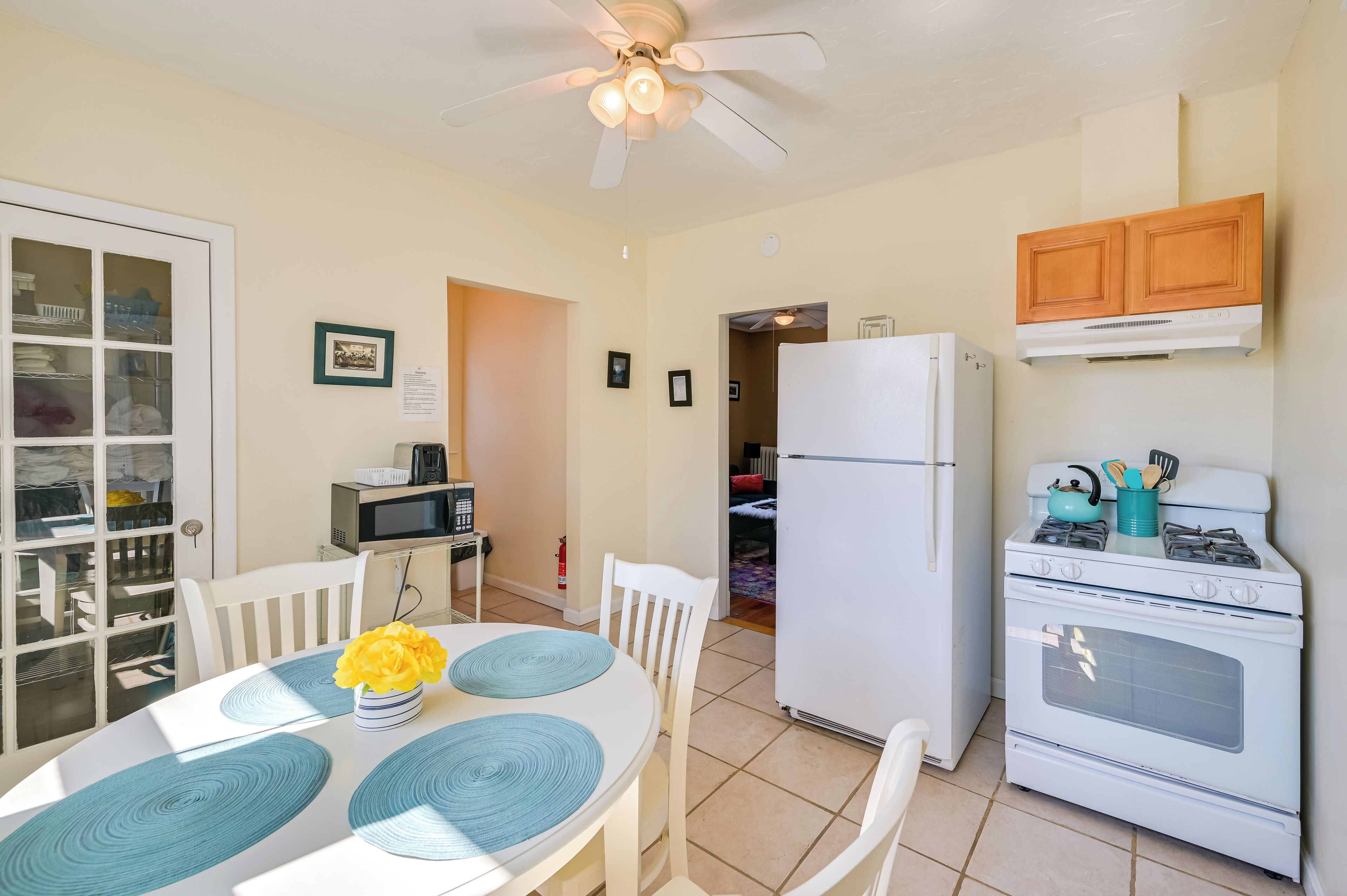 A compact kitchen with a white stove, a microwave, and a refrigerator. There is a dining table with blue placemats and a vase with yellow flowers.