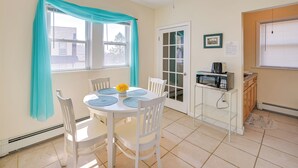Un comedor con una mesa y sillas blancas, una ventana con cortinas color turquesa, una zona de cocina con microondas, y una puerta que da al exterior.
