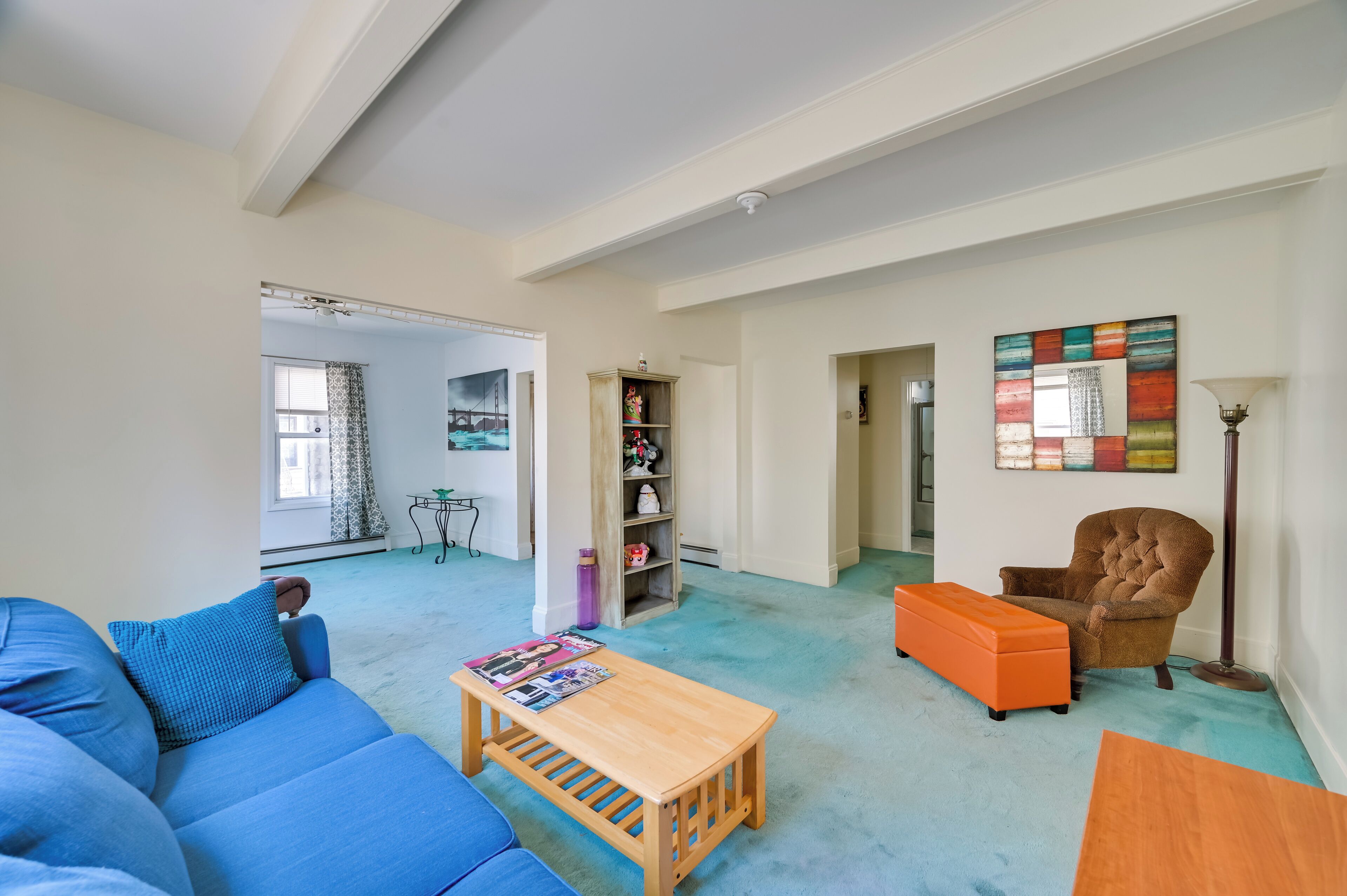 Een ruime woonkamer met een blauwe bank, een oranje poef en een houten salontafel. Er staat een boekenkast met diverse spullen in en aan de muur hangt een kleurrijk abstract schilderij.