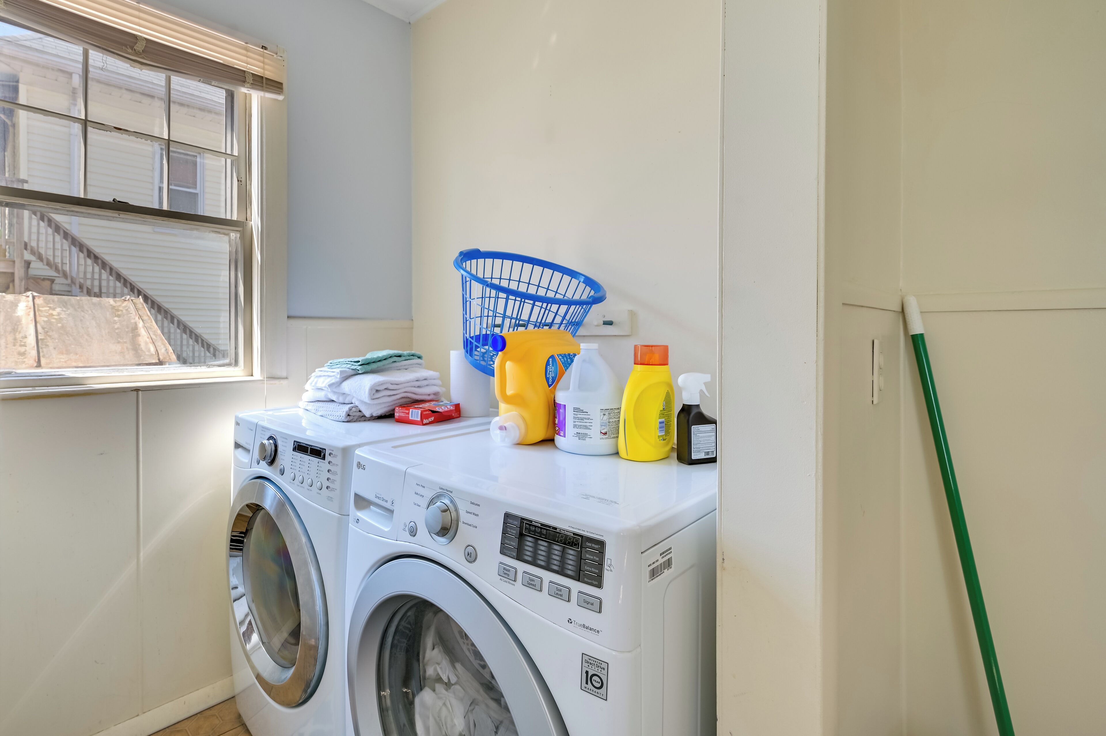 A laundry area with a front-loading washing machine, a blue laundry basket, and various cleaning supplies on top.