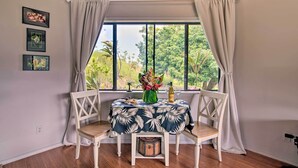 A dining table with a floral tablecloth, two white chairs, a vase with flowers, a bottle, and a wine glass on a window seat with curtains. - Hilo Couples Retreat ~ 6 Miles to Honoli'i Beach! (Hilo)