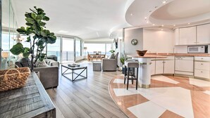 A modern living room with a large plant, a dining area, and a kitchen with white cabinets and a built-in oven.