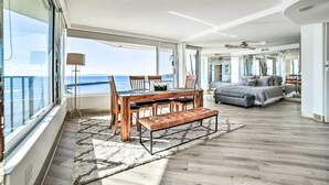 A spacious room with a large dining table, a sofa, and a bench. There is a large window offering a view of the ocean.