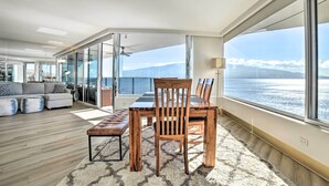 A modern living room with a wooden dining table and chairs, a sofa set, and a large window offering a view of the sea and mountains.