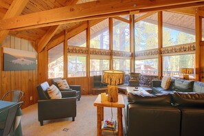 L’intérieur chaleureux d’un chalet avec une cheminée, des meubles en cuir et de grandes fenêtres donnant sur la forêt environnante.