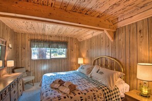 Un dormitorio de cabaña de madera con una cama, dos lámparas de noche, un tocador y una ventana con cortina.
