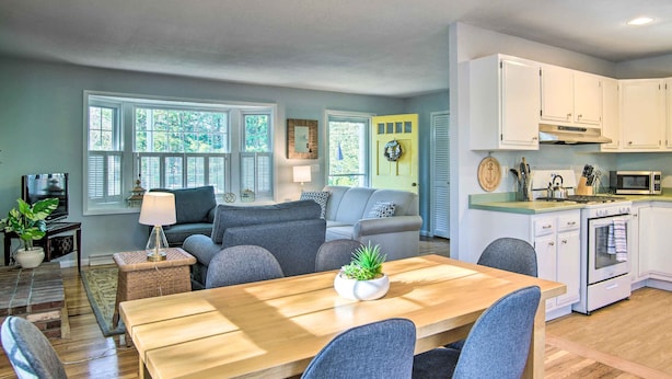 A modern kitchen with white cabinets, a microwave, stove, and a dining area with a wooden table and chairs. - Hen Cove Home w/ Wood Deck - Walk to Beaches! (Cataumet)