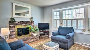 A living room with a fireplace, a television, and a large window. - Hen Cove Home w/ Wood Deck - Walk to Beaches! (Cataumet)