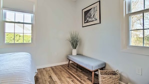 A bedroom with a bed, a bench, a potted plant, a framed picture, and a wicker basket. - Hen Cove Home w/ Wood Deck - Walk to Beaches! (Cataumet)