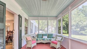 A covered porch with wicker chairs and a bench, large windows, and a view of the outdoors. - Charming Nineteenth-century Cape Code Escape (Centerville)