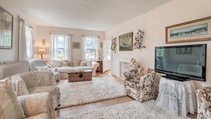 A cozy living room with a fireplace, a television, and a large window with curtains. - Charming Nineteenth-century Cape Code Escape (Centerville)