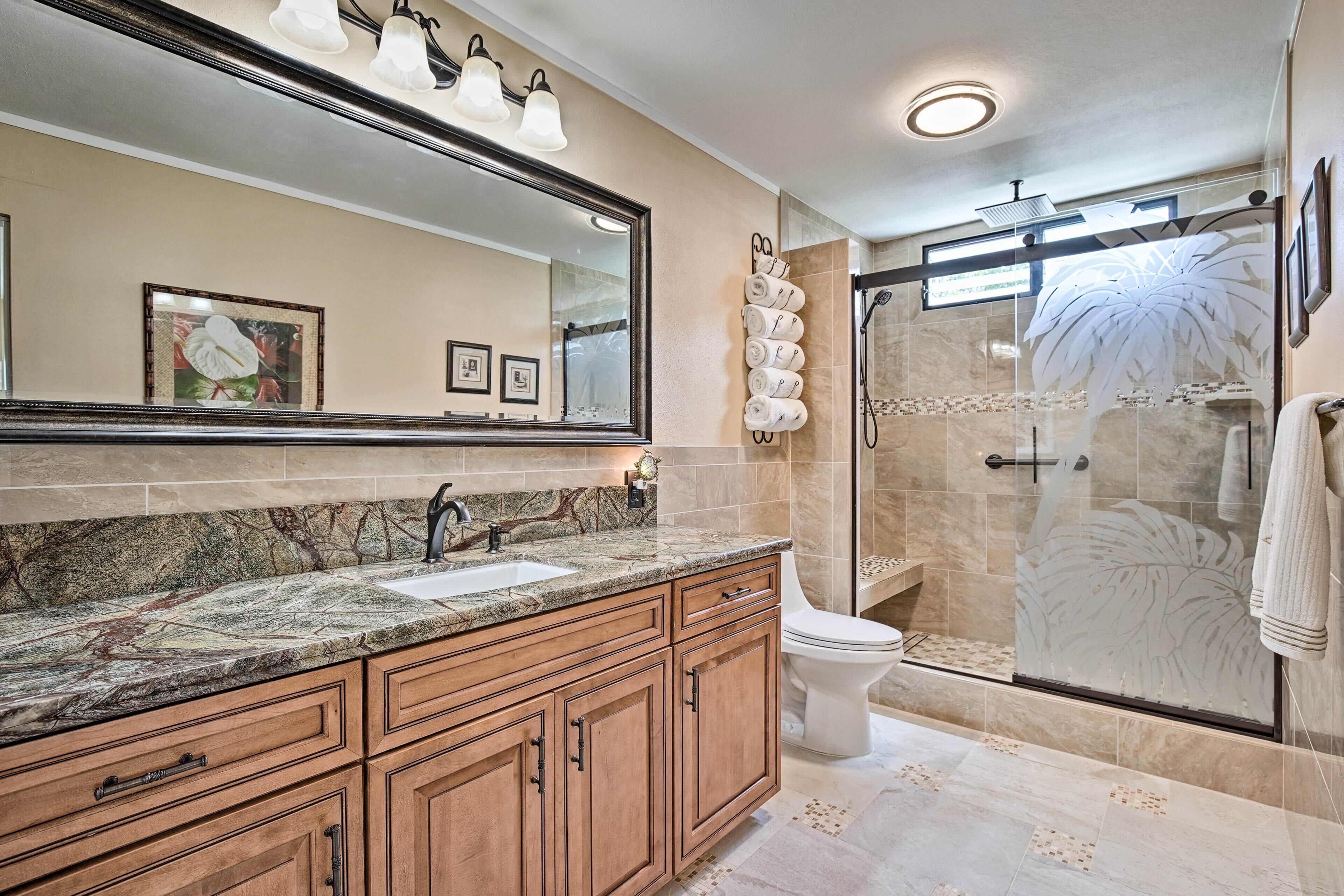 A bathroom with a marble countertop, wooden cabinets, a large mirror, and a glass shower enclosure.