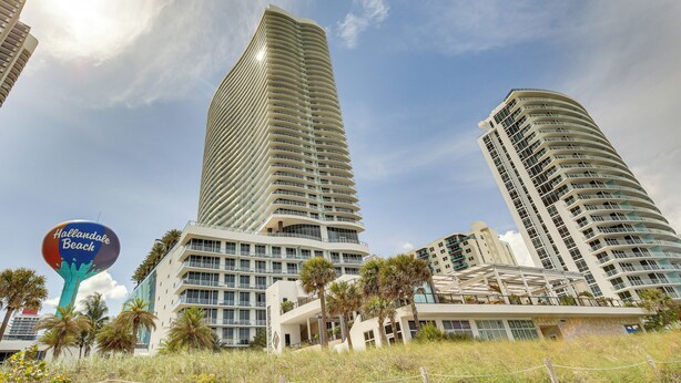 Un hôtel moderne à la façade incurvée, avec une enseigne bien visible indiquant « Hollandale Beach », et des palmiers au premier plan.