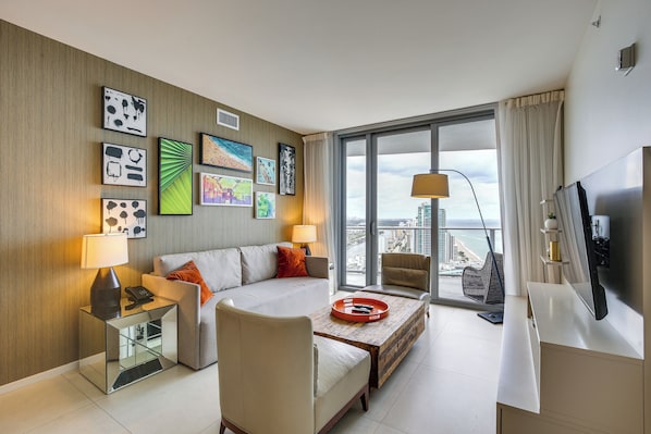 A modern hotel room with a sofa, armchair, and coffee table. There are artworks on the wall, a TV, and a view of the city. - Luxe Hollywood Condo: Resort Perks + Beach Access! (Hollywood)