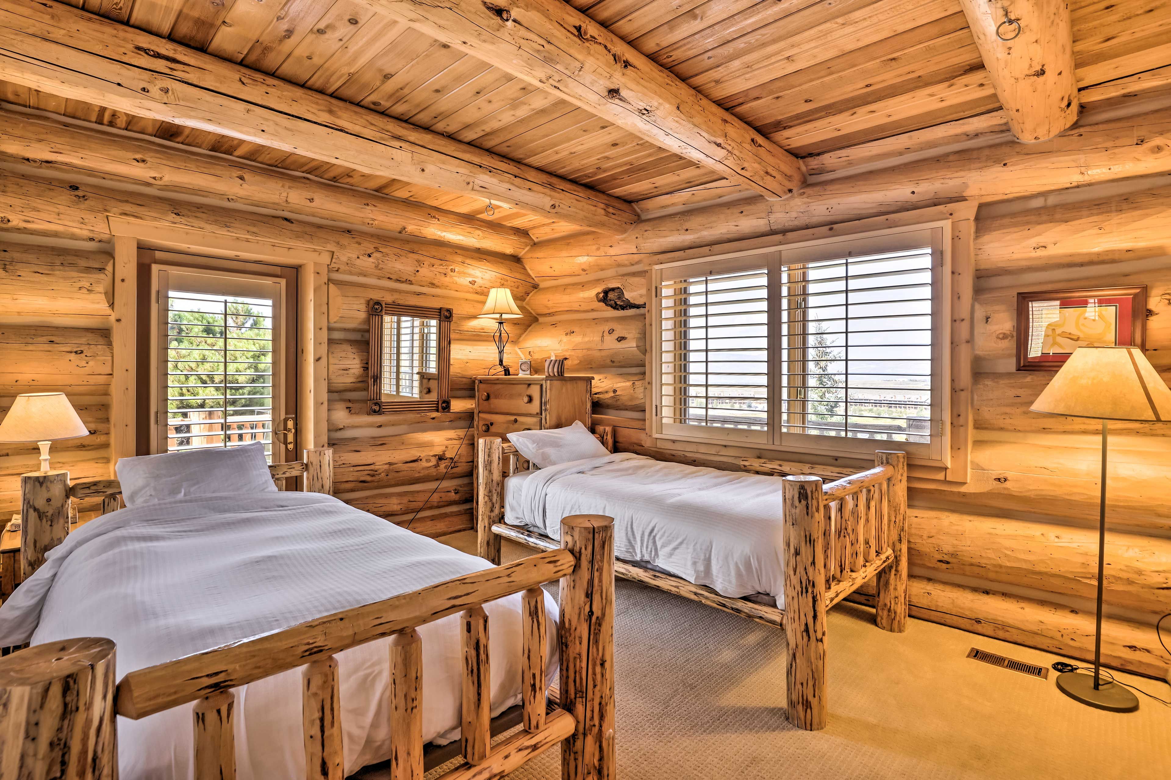 Quarto de cabana de madeira com duas camas de madeira, janela com persianas, abajur e quadro na parede.