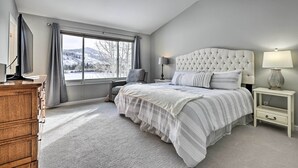 Une chambre avec une grande fenêtre, un lit doté d’une tête de lit capitonnée, une table de chevet avec une lampe et une commode en bois.