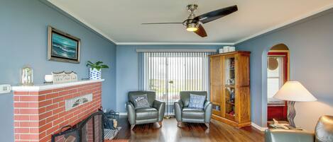 A living room with a fireplace, two armchairs, a sofa, a ceiling fan, and a large window with blinds.