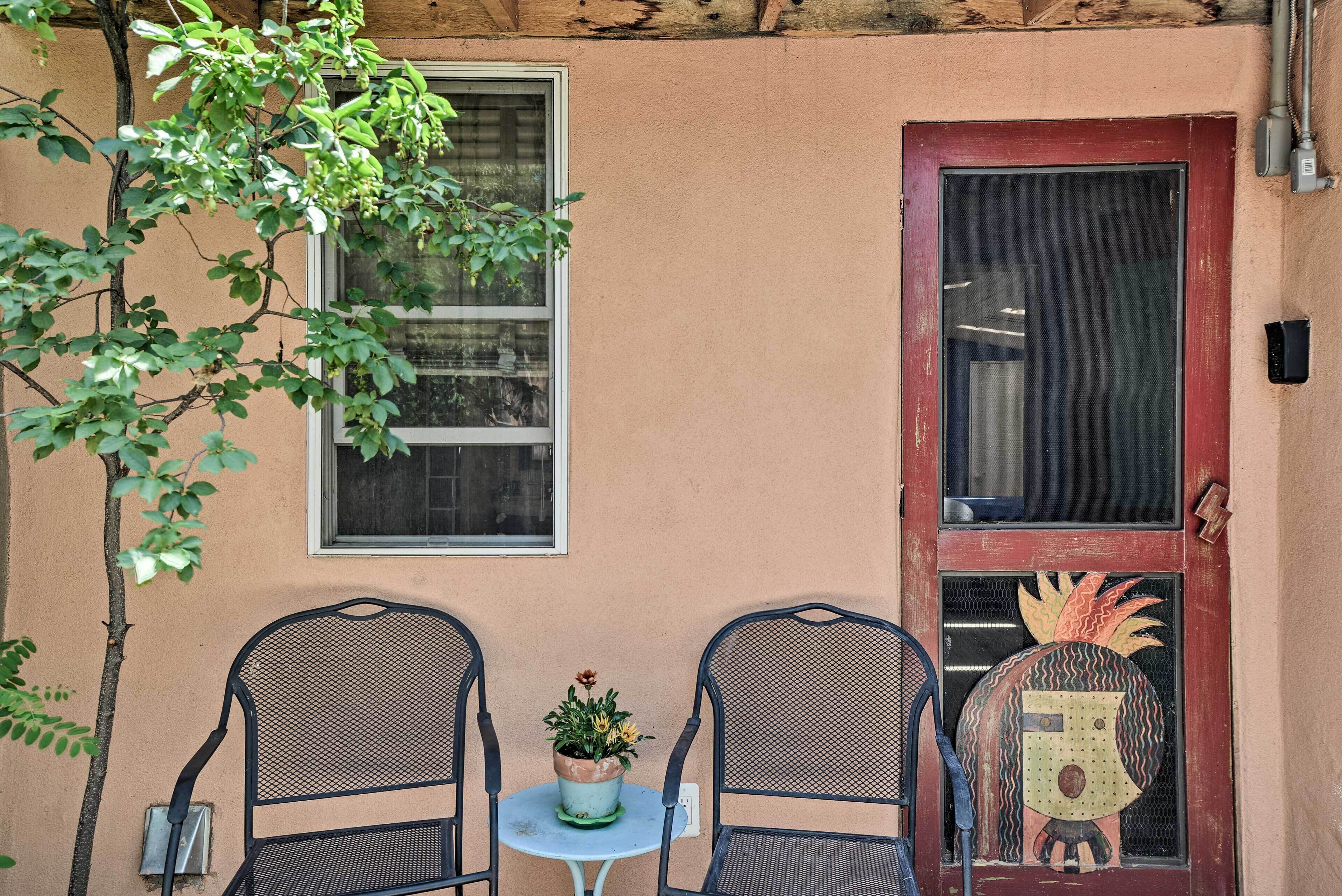 Un petit espace extérieur aménagé avec deux chaises en métal et une table, un pot de fleurs et une porte ornée d’un motif décoratif.