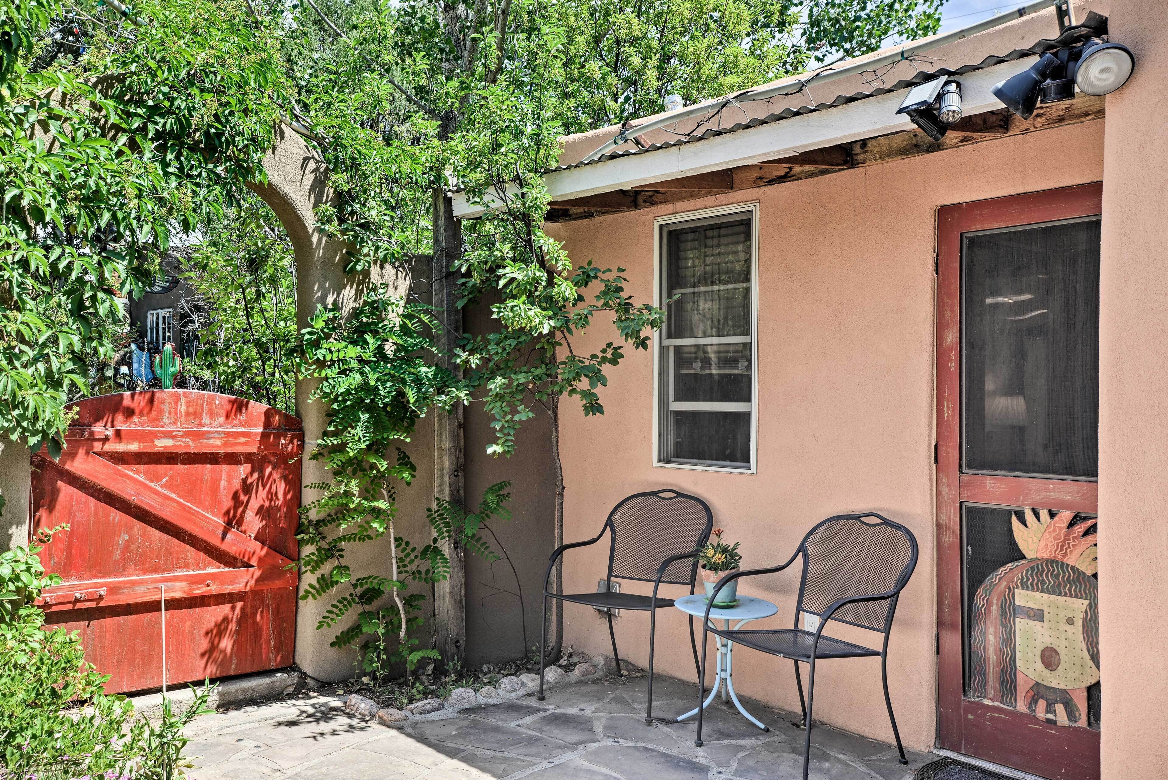 A small outdoor seating area with two chairs and a table, a red gate, and a window with a decorative painting.