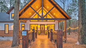 A cabin with a wooden deck leading to a covered porch, surrounded by trees. - Modern Cabin w/ Hot Tub: 1 Mi From Hochatown! (Broken Bow)