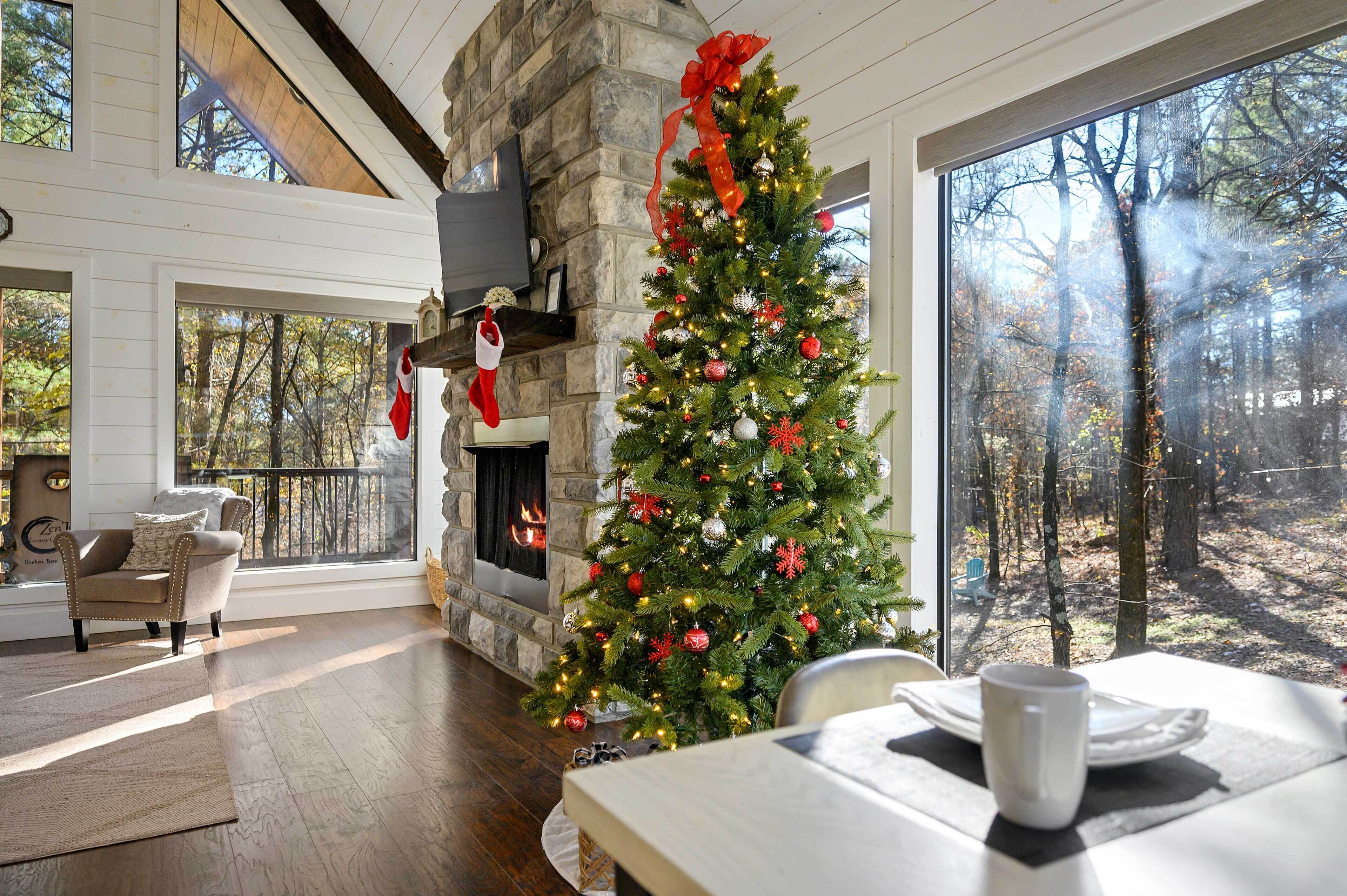 A cozy living room with a stone fireplace, a Christmas tree, and a large window overlooking a forest.
