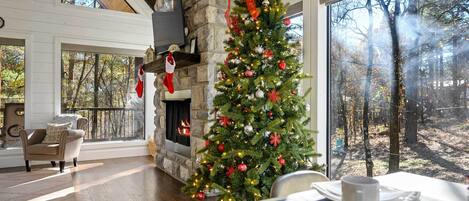 A cozy living room with a stone fireplace, a Christmas tree, and a large window overlooking a forest.