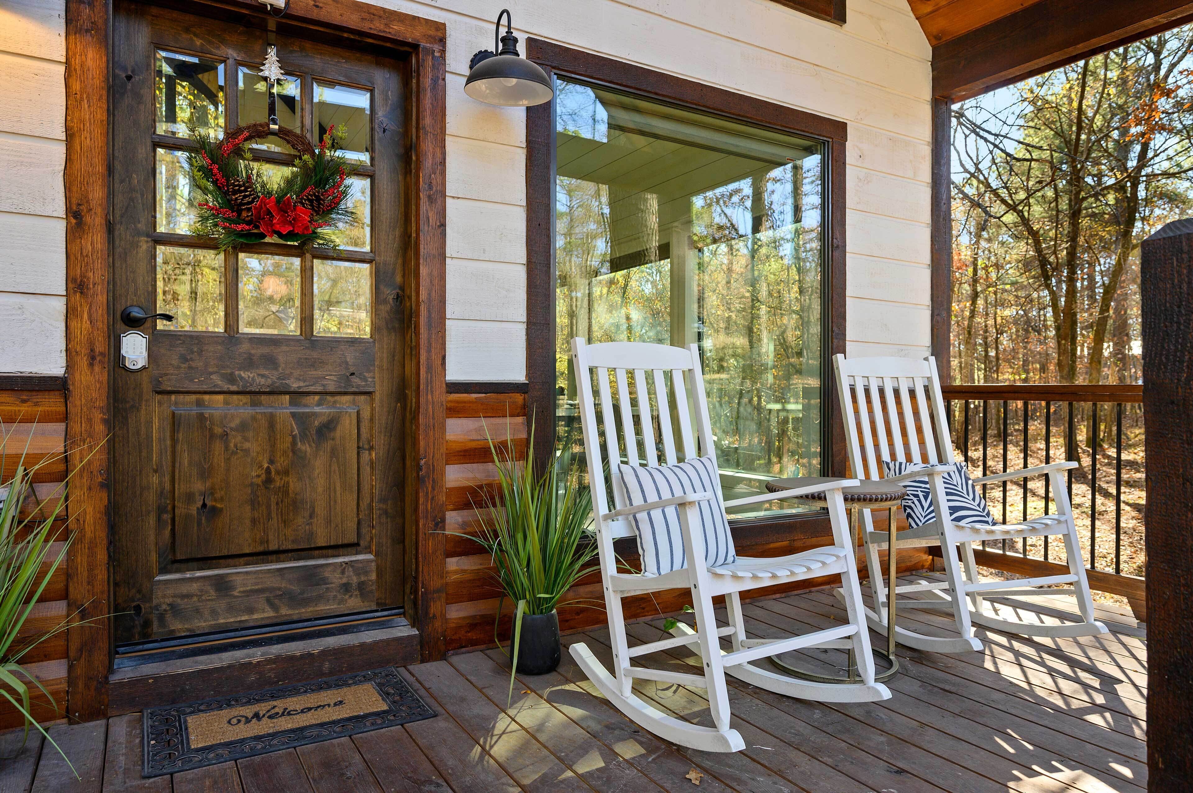 A wooden door with a wreath, a porch with two rocking chairs, and a large window overlooking trees.