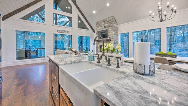 A spacious kitchen with a large island, stone fireplace, and large windows offering views of the outdoors. - Modern Cabin w/ Hot Tub: 1 Mi From Hochatown! (Broken Bow)