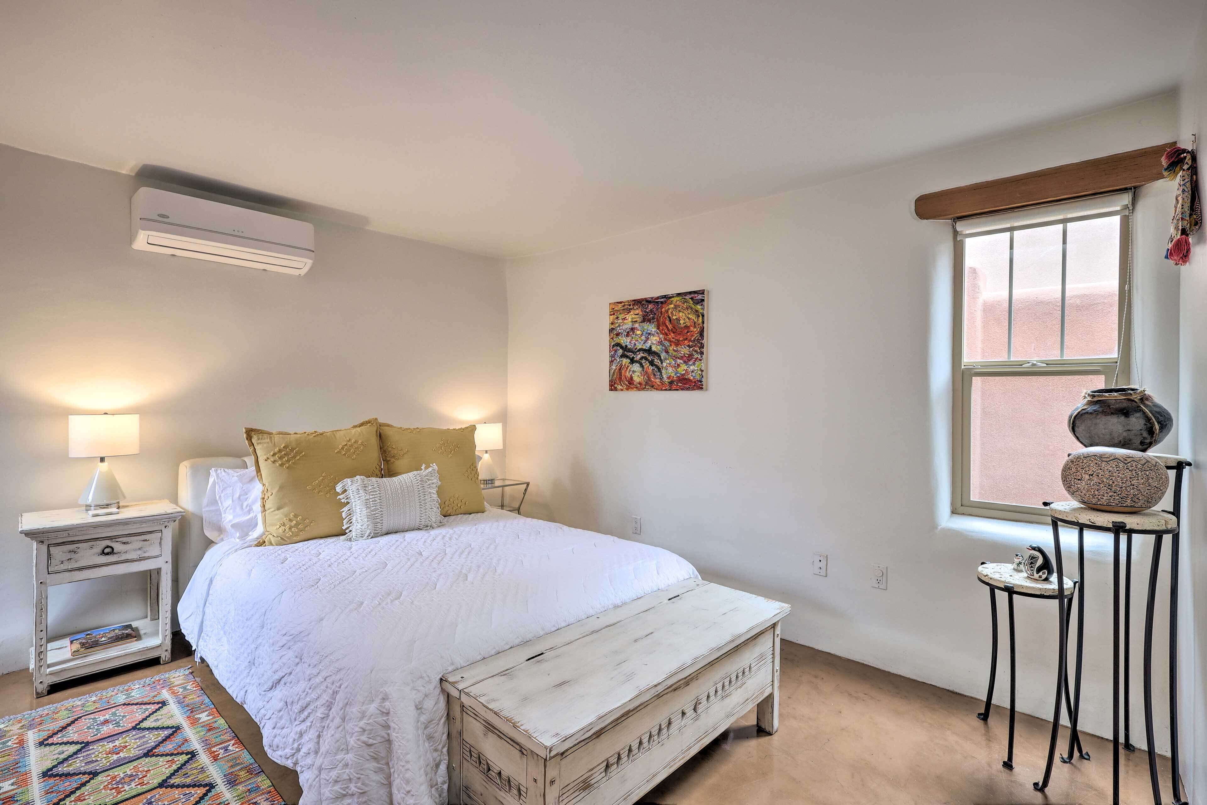 A bedroom with a bed, bedside tables, a bench, a window, and a wall-mounted air conditioner.